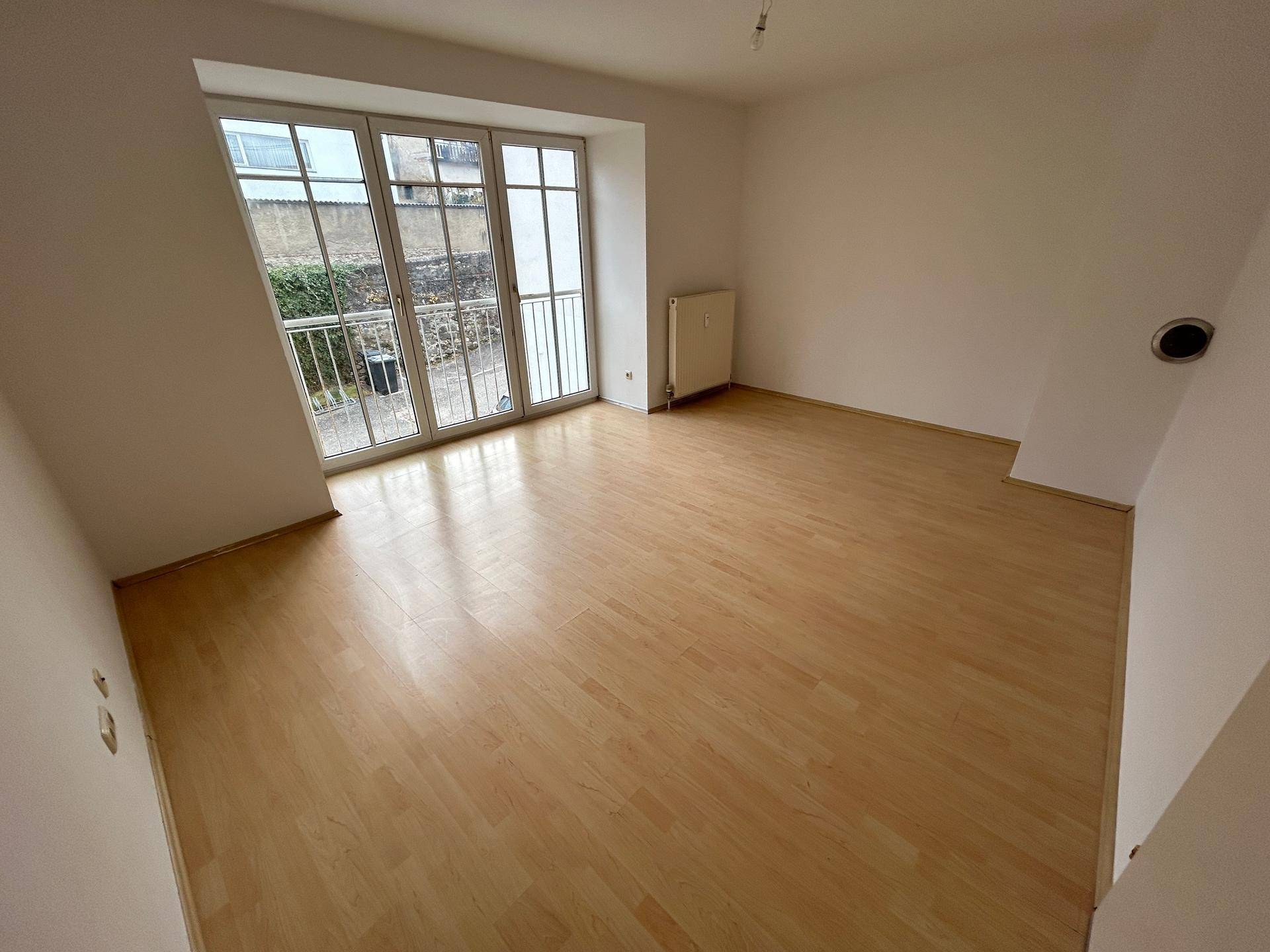 Heller Raum mit großen Fenstern, französischem Balkon und hellem Laminatboden, ideal als Wohnzimmer.