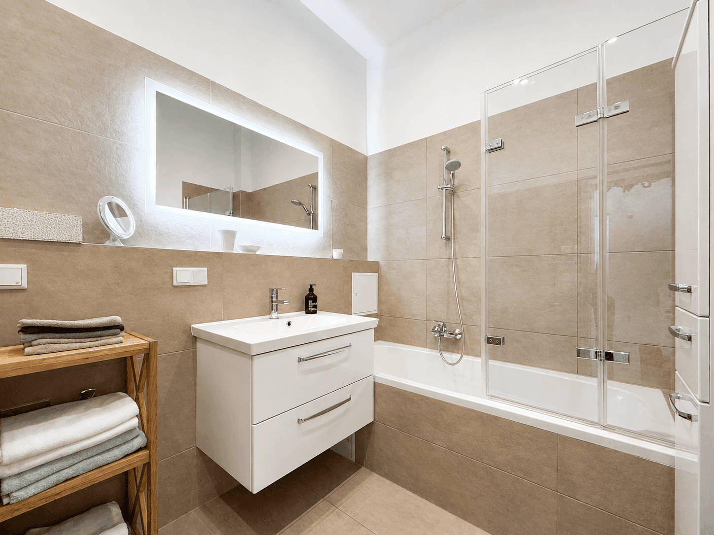 Modernes Badezimmer mit beigefarbenen Fliesen, Badewanne mit Dusche und beleuchtetem Spiegel.