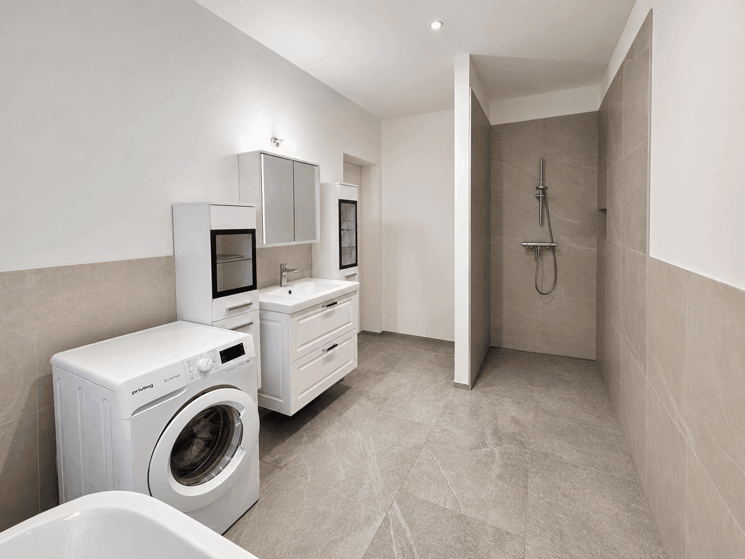 Funktionales Badezimmer mit Waschmaschine, Waschtisch und ebenerdiger Dusche in hellen Farbtönen.