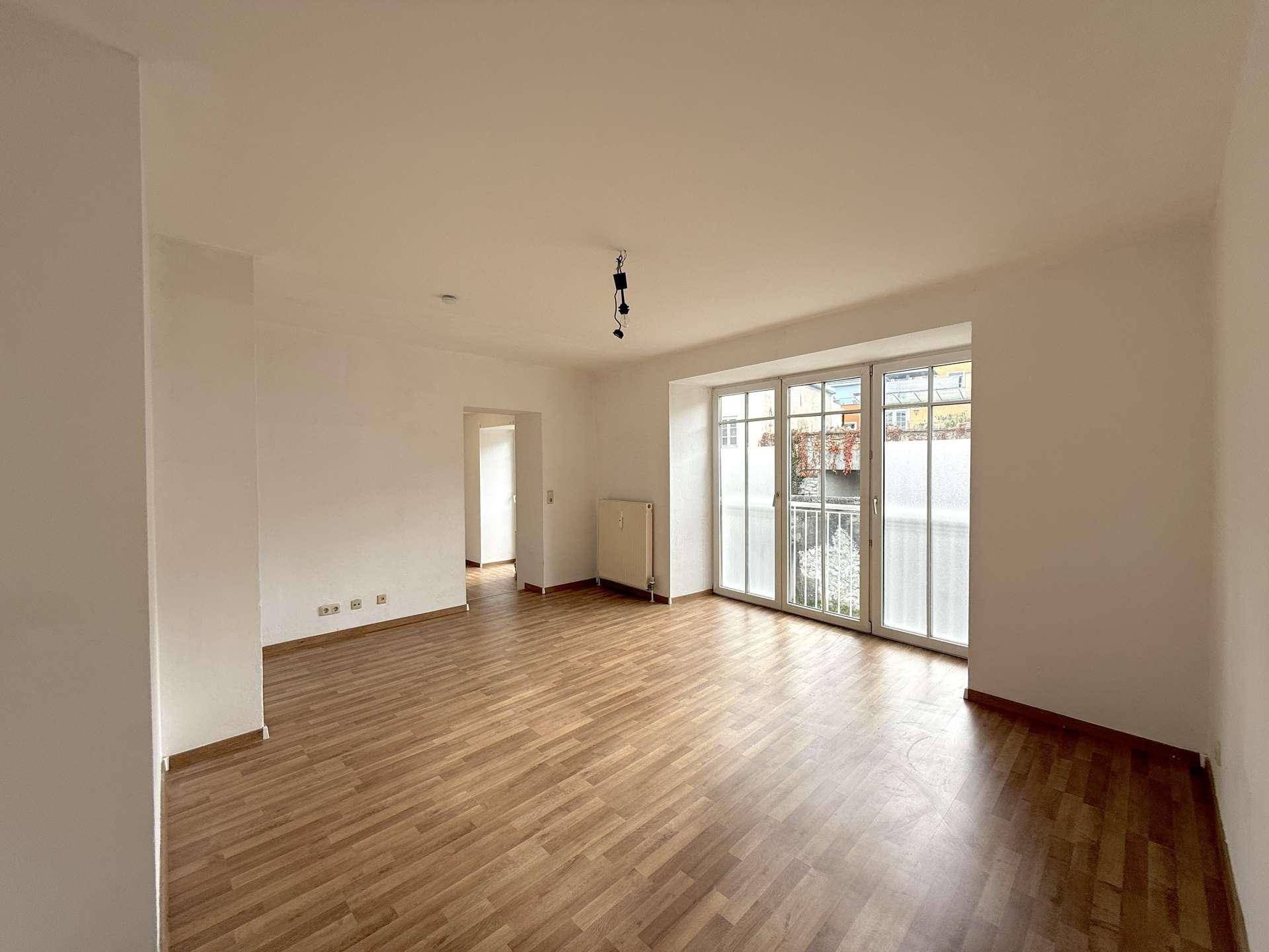 Geräumiges, helles Wohnzimmer mit Laminatboden, Heizkörper und Zugang zu einem weiteren Raum.