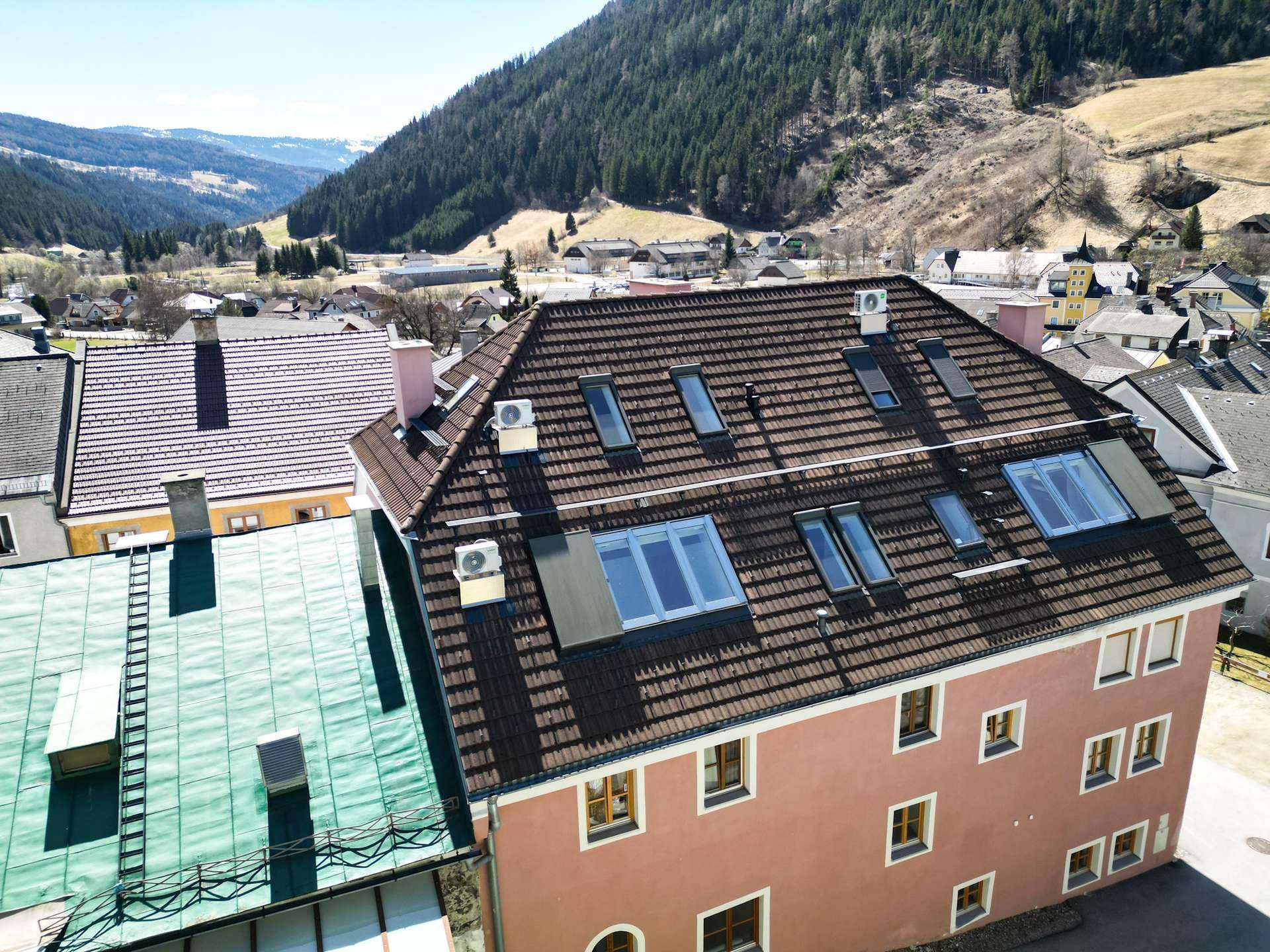 Dach eines Gebäudes mit zahlreichen Dachfenstern und Klimaanlagen in einer bergigen Umgebung.