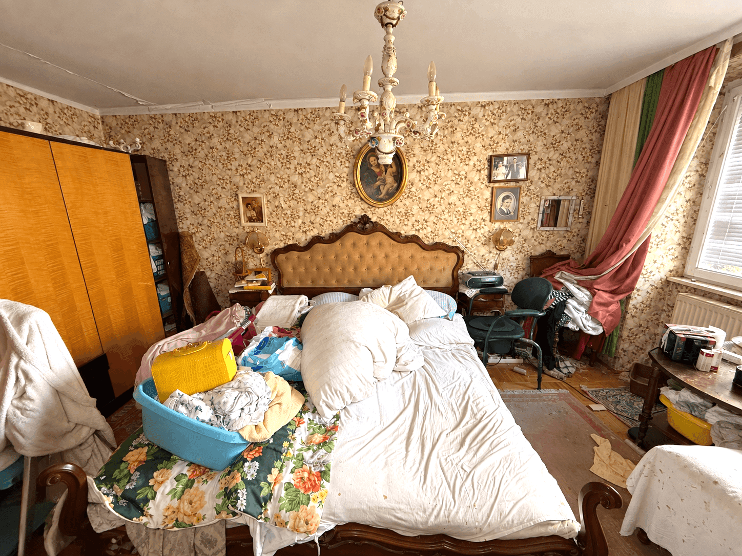 Schlafzimmer mit großem Bett, floraler Tapete und Kronleuchter, viele Gegenstände.