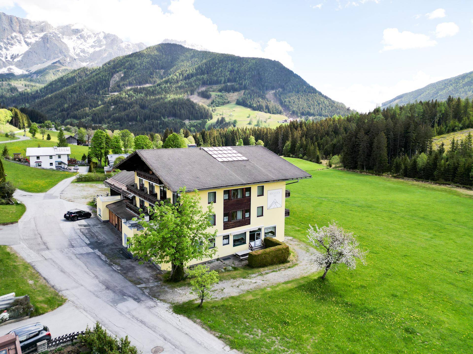 Gelbes Gebäude mit Solarpaneelen auf dem Dach, umgeben von einer grünen Wiese und bewaldeten Bergen.