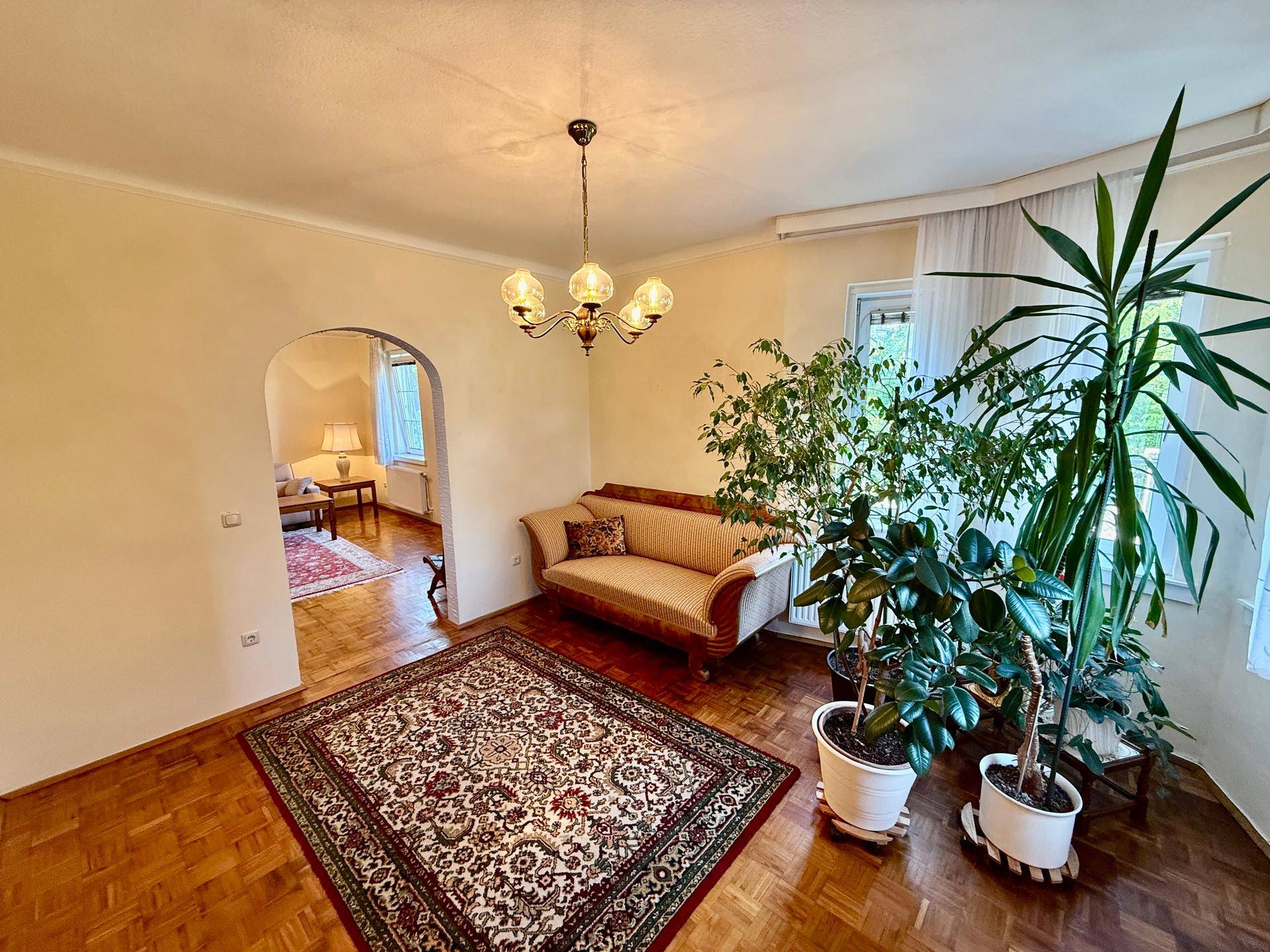 Helles Wohnzimmer mit Parkettboden, klassischem Sofa und großen Zimmerpflanzen.