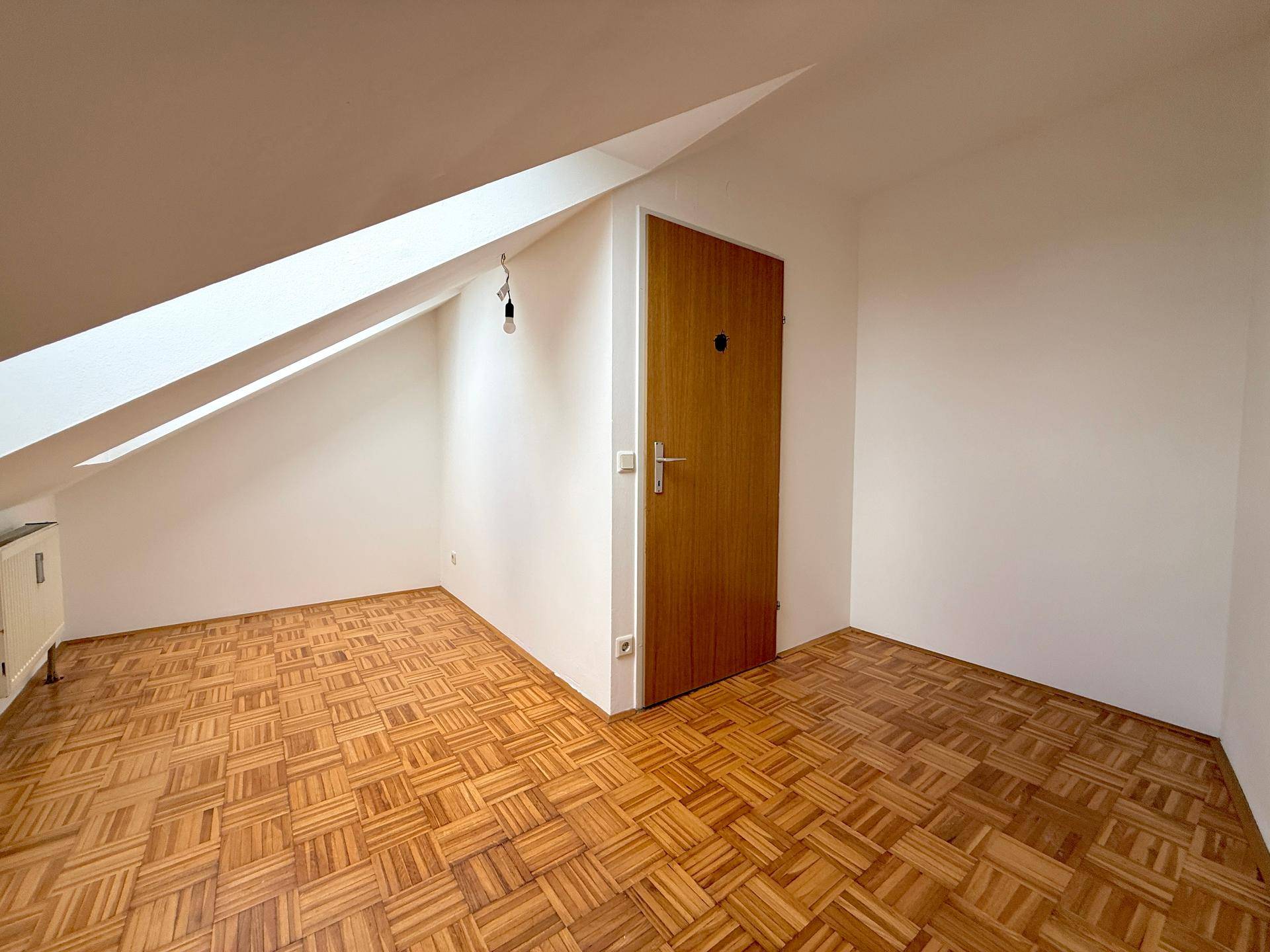 Leerer Raum im Dachgeschoss mit schrägen Wänden und Parkettboden.