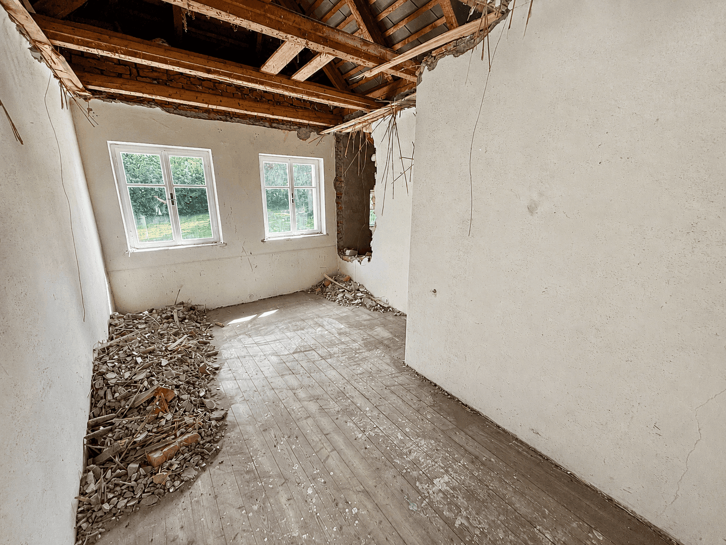 Dachgeschossraum im Umbau mit freigelegten Holzbalken, Schutt und zwei Fenstern.
