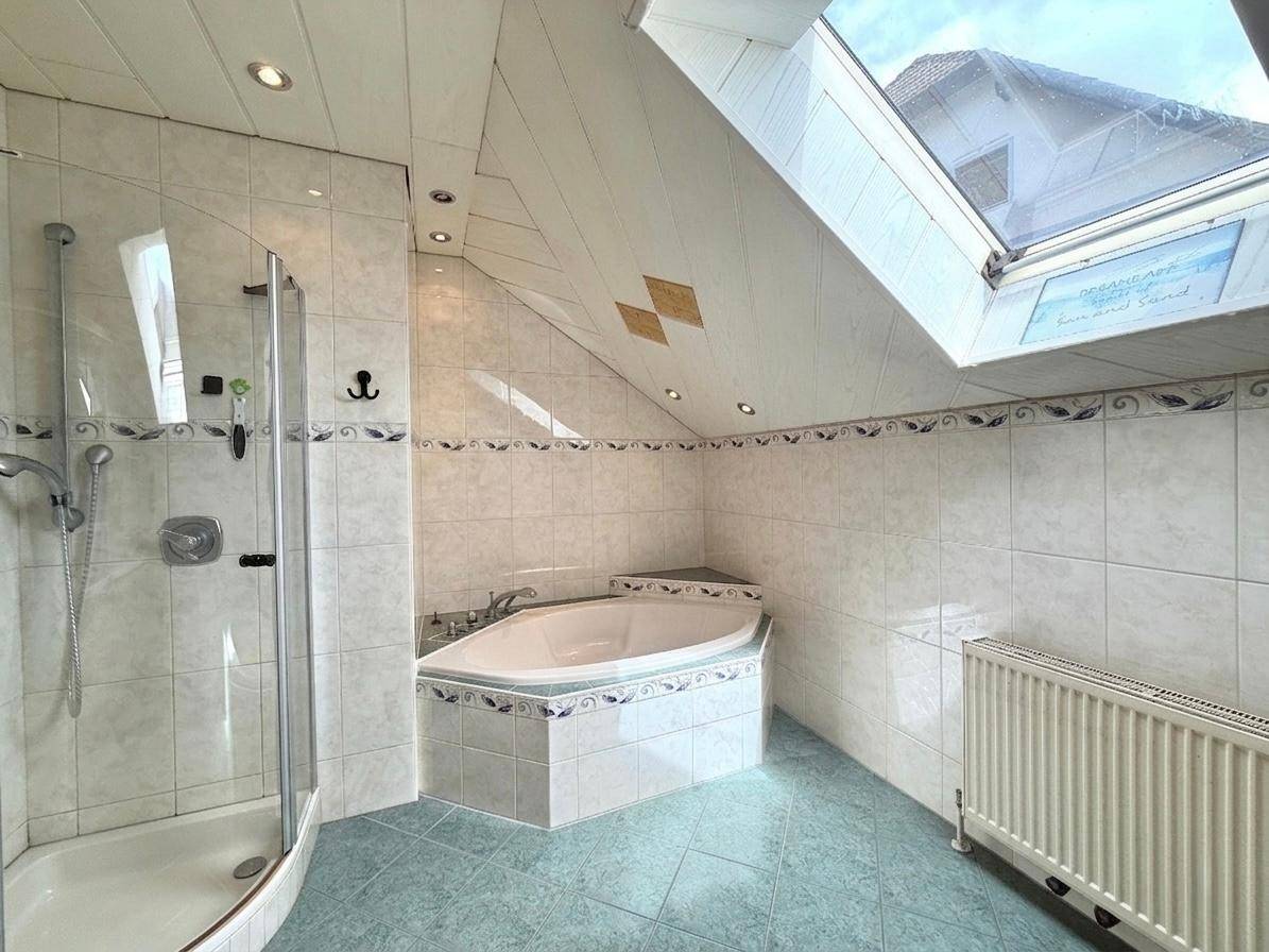 Badezimmer im Dachgeschoss mit Eckbadewanne, Duschkabine, Dachfenster und hellen Fliesen.