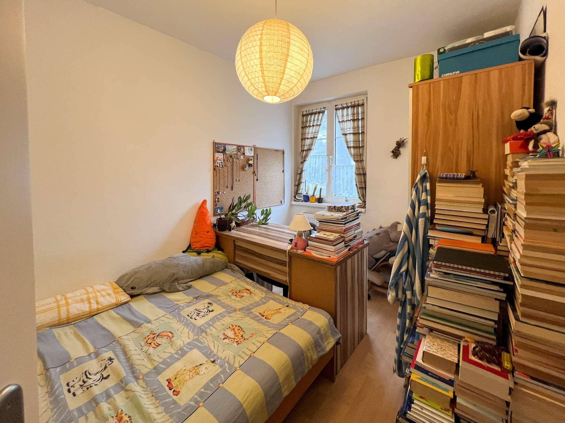 Gemütliches Schlafzimmer mit Einzelbett, Schreibtisch und vielen Büchern, hell durch ein Fenster.