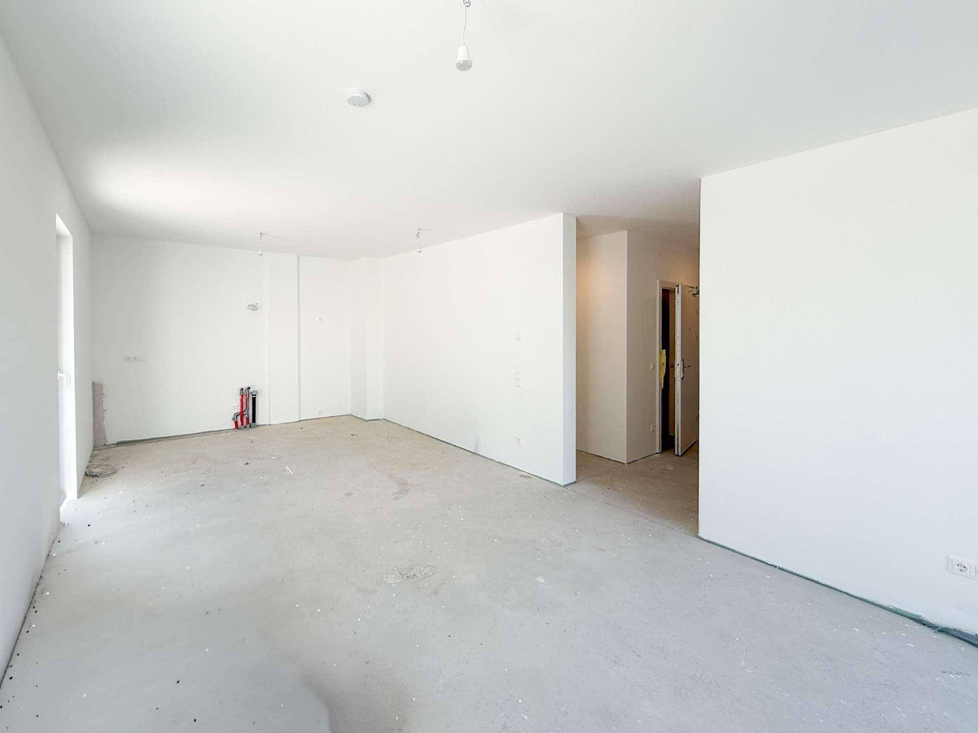 Geräumiger Rohbauraum mit Betonboden und weiß gestrichenen Wänden, offen zu einem weiteren Bereich.