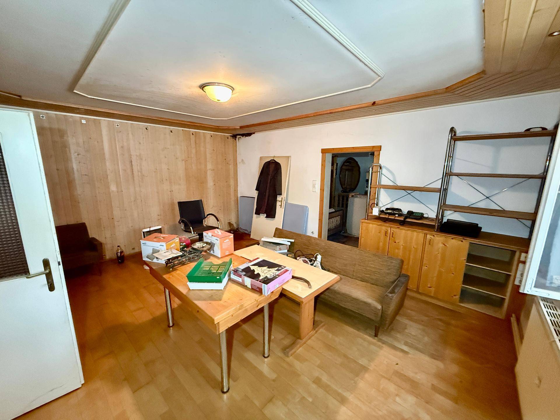 Wohnraum mit Holzboden, Holzvertäfelung und Deckenpaneelen, derzeit als Arbeitsbereich genutzt.