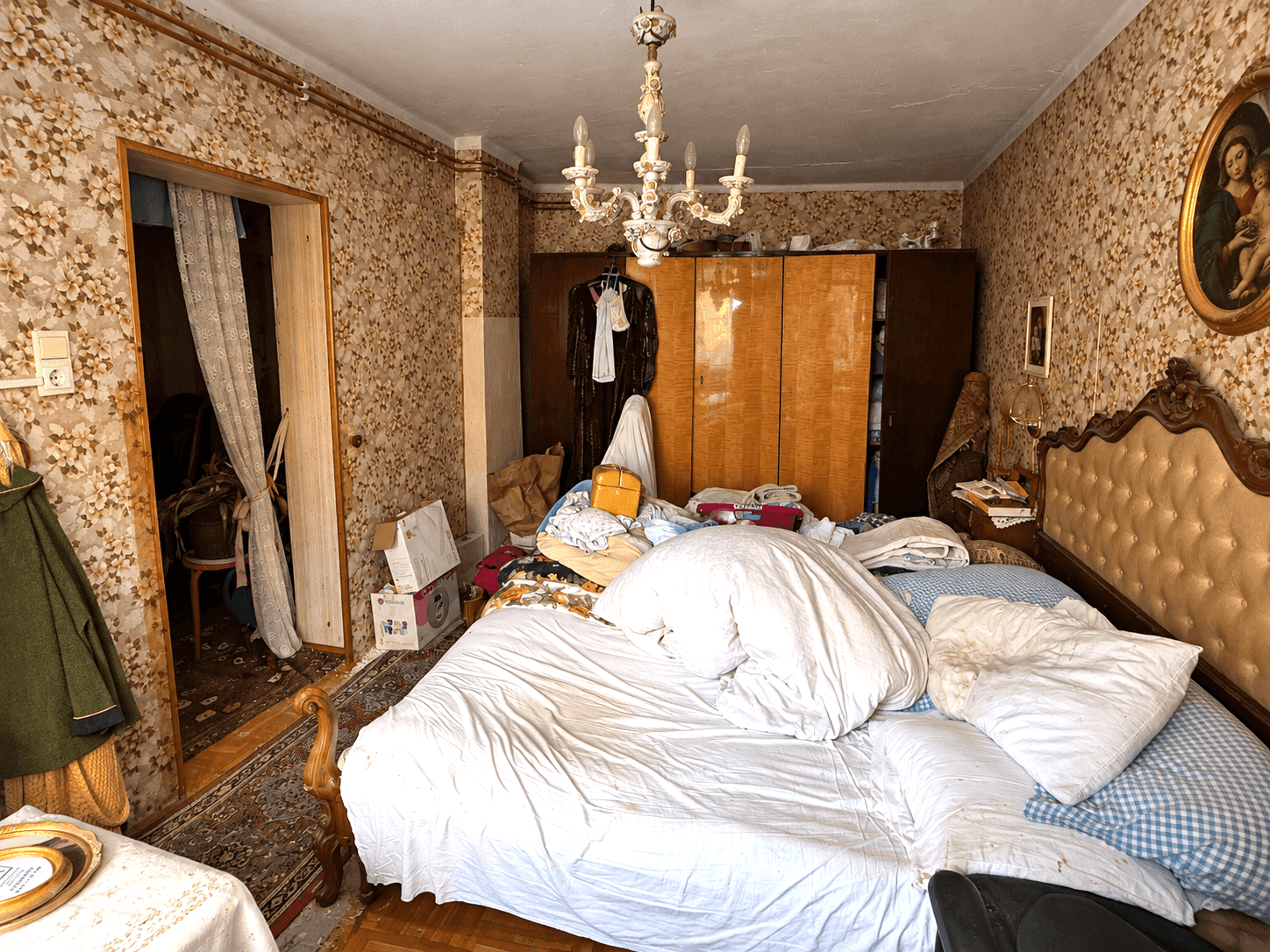 Schlafzimmer mit Doppelbett, floraler Tapete und Holzschränken, unaufgeräumt.