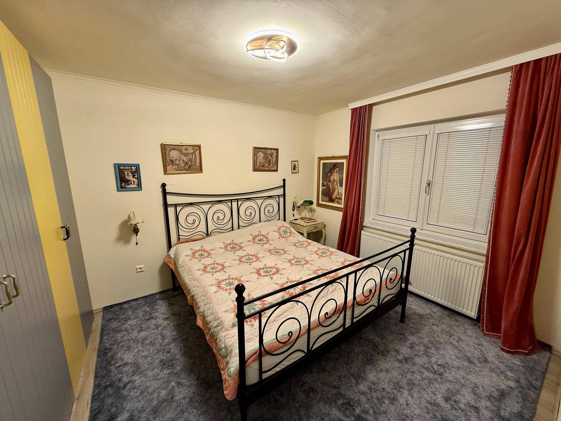 Schlafzimmer mit schmiedeeisernem Bett, Teppichboden und Fenster mit roten Vorhängen.