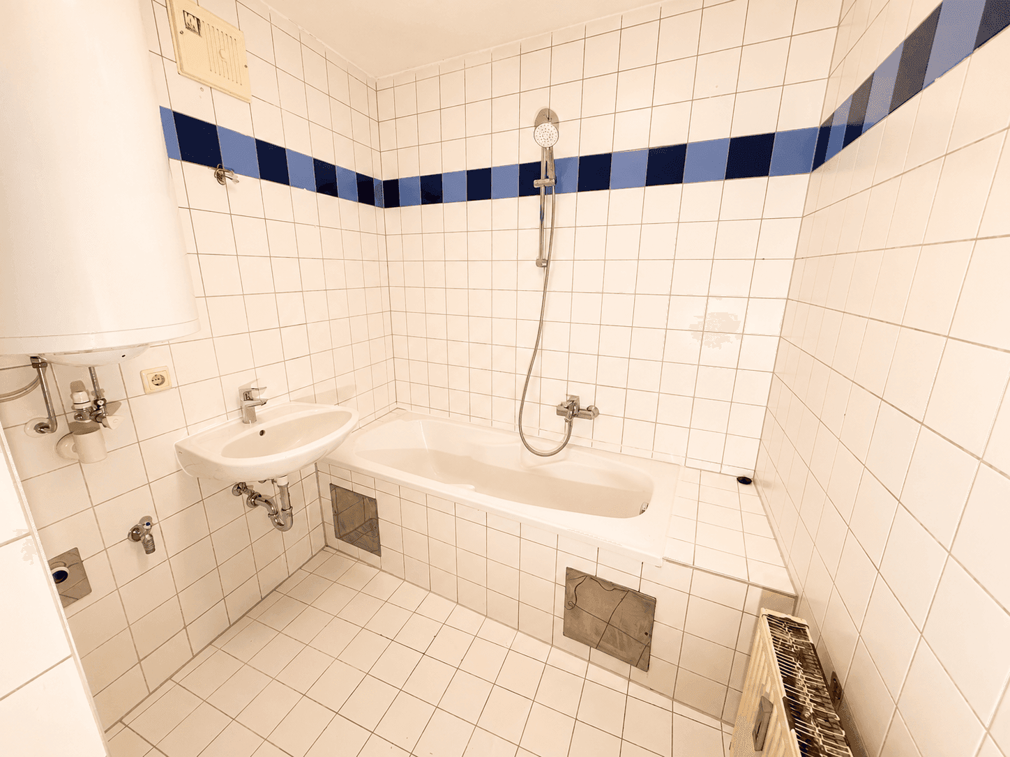 Badezimmer mit weißen Fliesen, Badewanne mit Duschfunktion, Waschbecken und einem blauen Dekorstreifen.