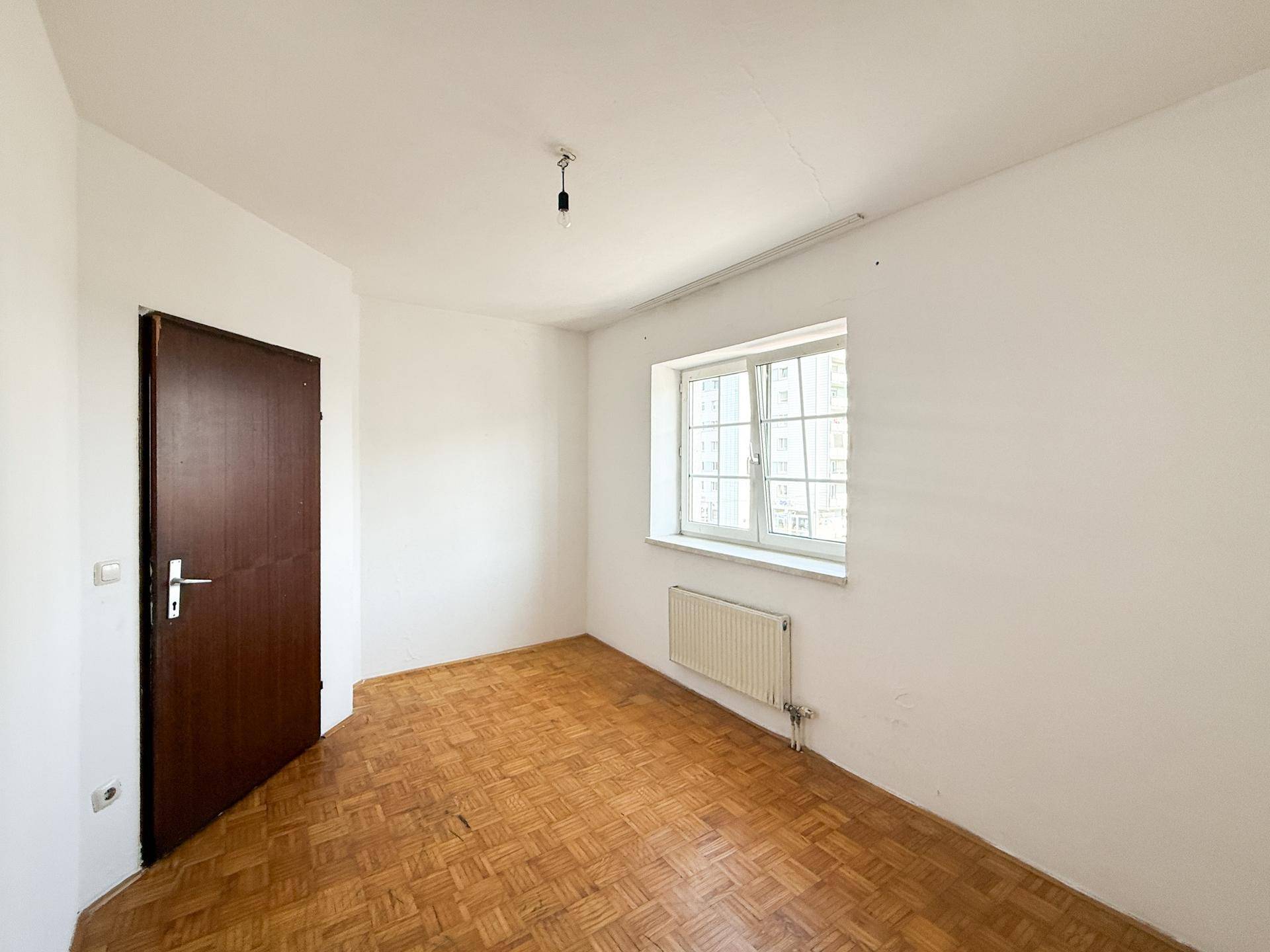 Helles Schlafzimmer mit Parkettboden und einem Fenster, das viel Tageslicht hereinlässt.