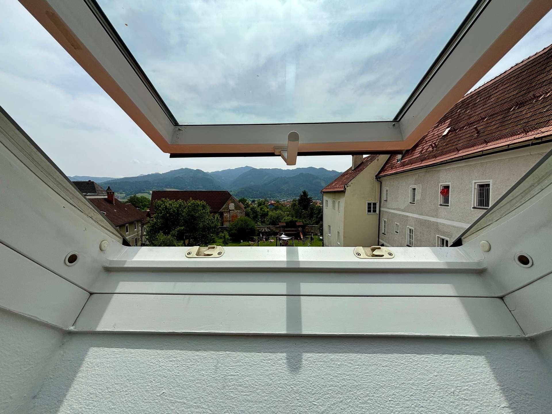 Blick aus dem Dachfenster auf die umliegende Landschaft mit Häusern und Bergen im Hintergrund.