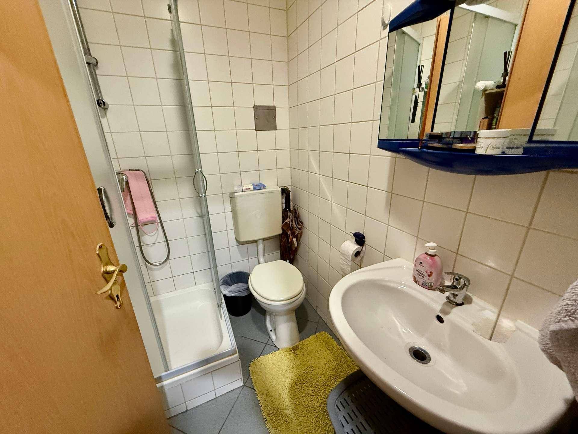 Kleines Badezimmer mit Duschkabine, WC und Waschbecken, gefliest in Weiß und Grau.