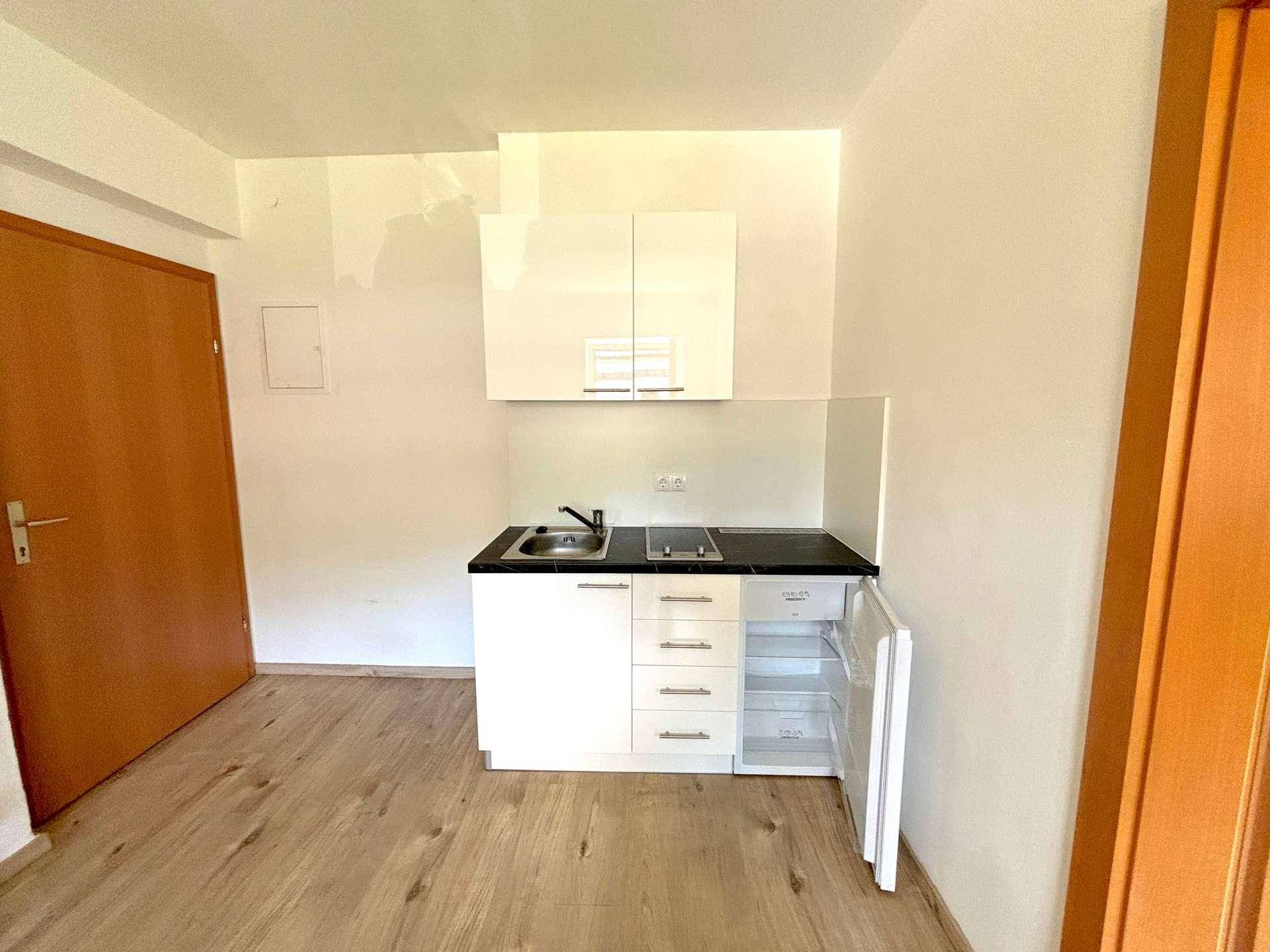Kompakte Küchenzeile mit weißen Hochglanzschränken, Spüle und integriertem Kühlschrank in einem hellen Raum.