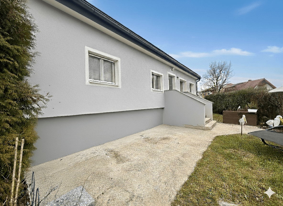 Seitenansicht des Hauses mit grauem Putz, kleinem Gartenbereich und Zugang zum Eingang.