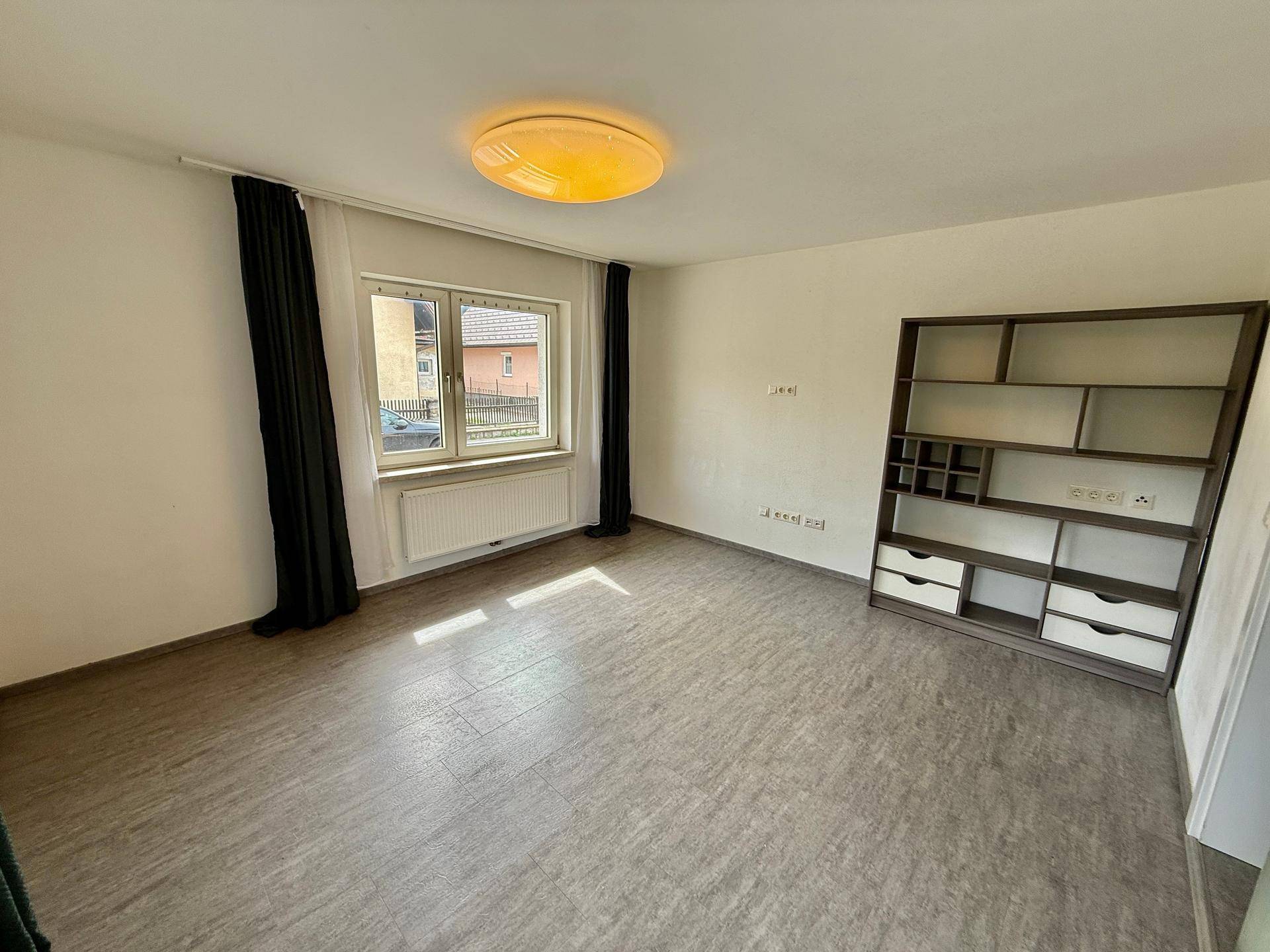 Helles Wohnzimmer mit Vinylboden, einem Fenster und einem Regal an der Wand.
