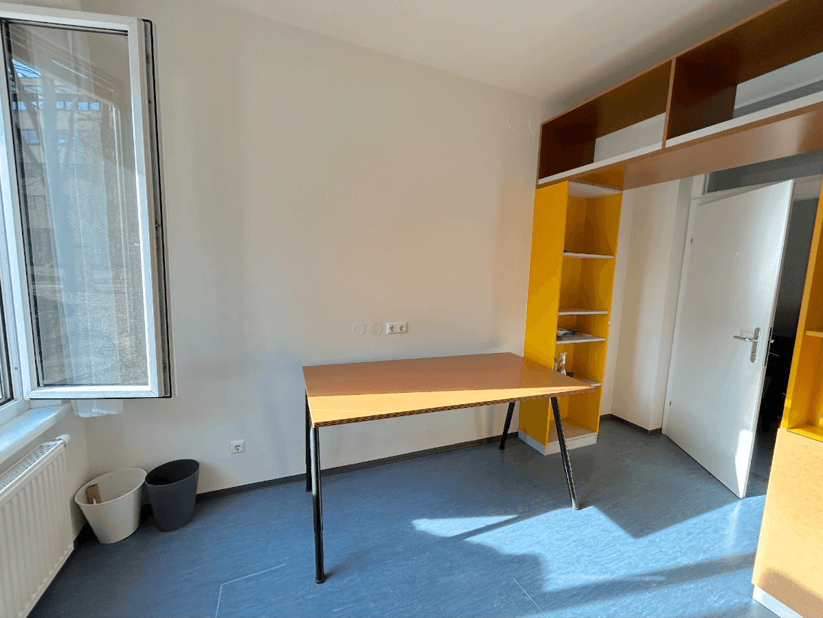 Helles Schlafzimmer mit großem Fenster, Schreibtisch und gelben Regalen für Bücher.