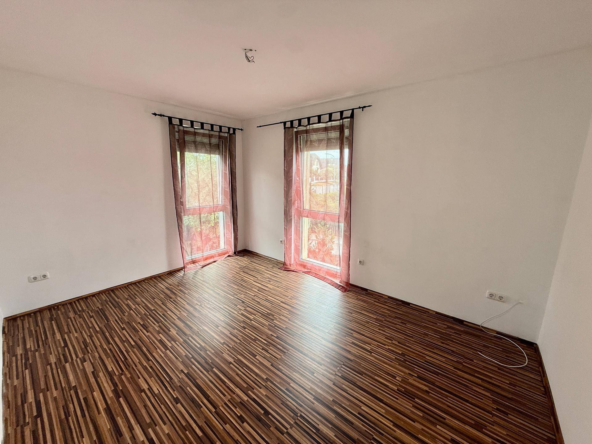 Helles Schlafzimmer mit Laminatboden und zwei Fenstern, geschmückt mit roten Vorhängen.