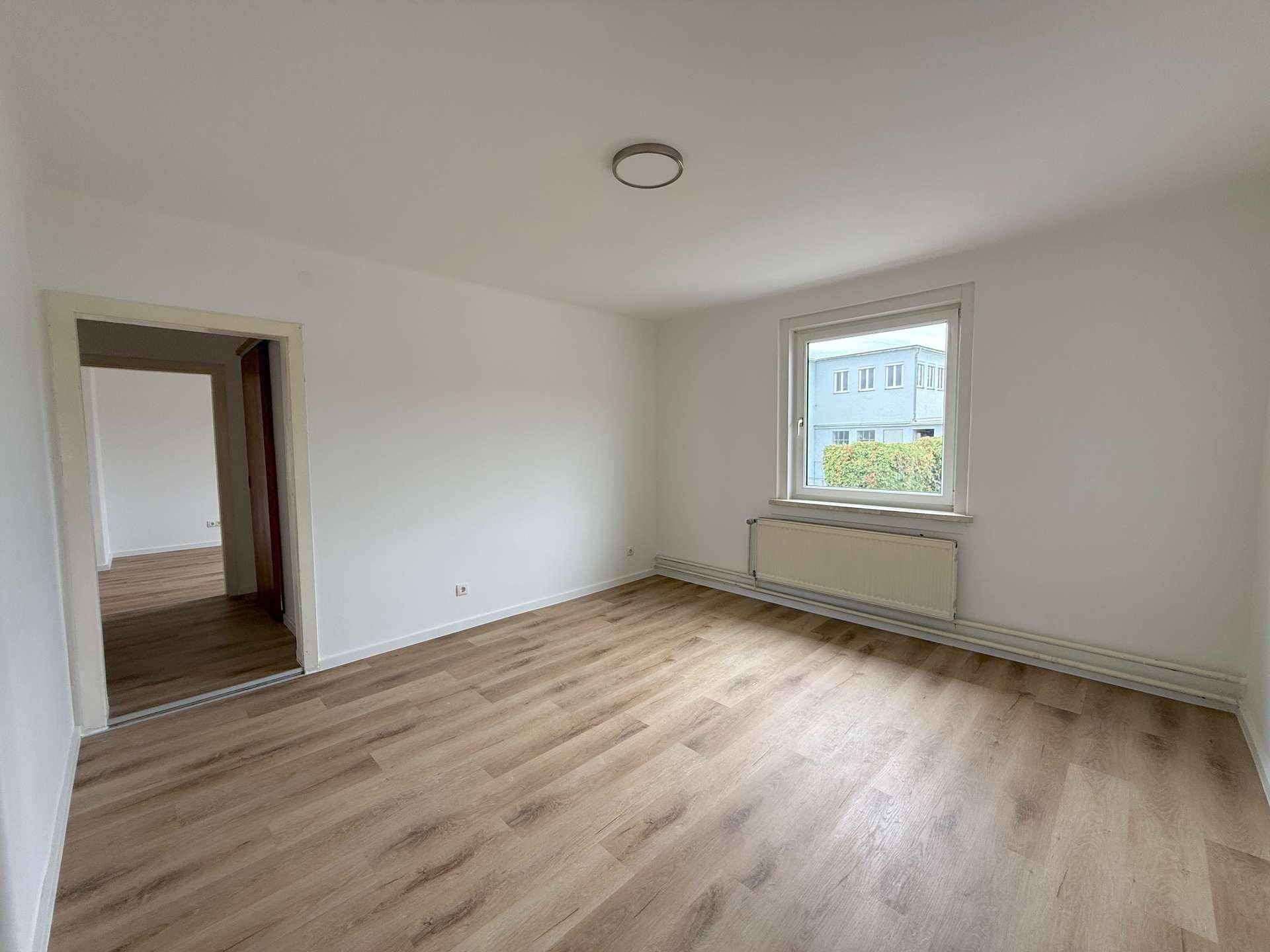 Geräumiges Zimmer mit hellem Laminatboden, weißen Wänden und Blick auf ein Gebäude.