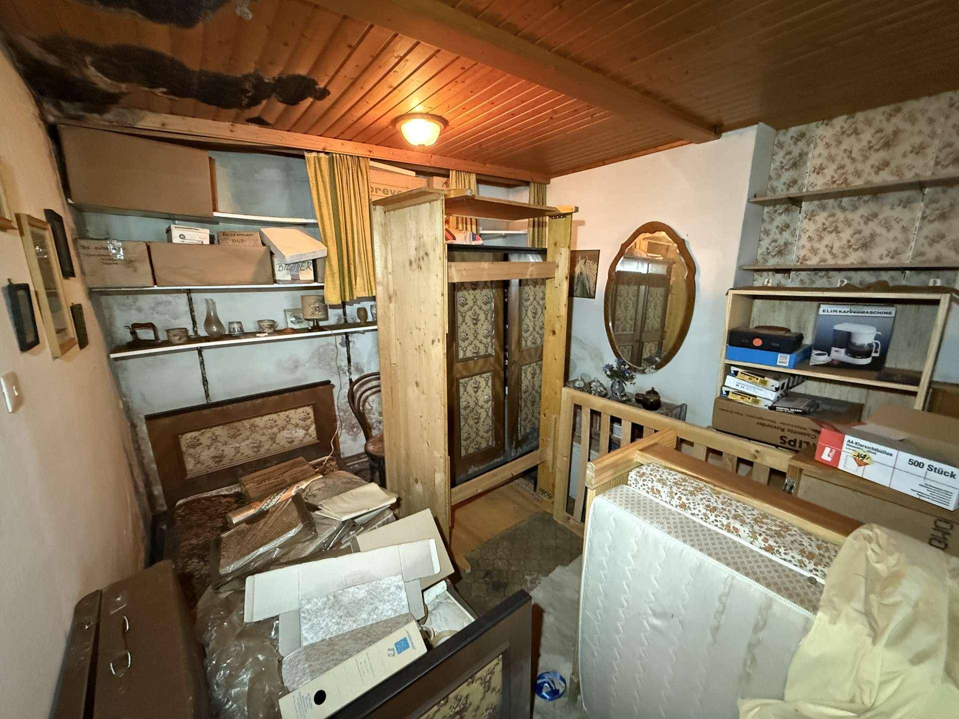 Überfülltes Schlafzimmer mit Holzdecke, Teppichboden und Vintage-Möbeln, die auf eine Renovierung warten.