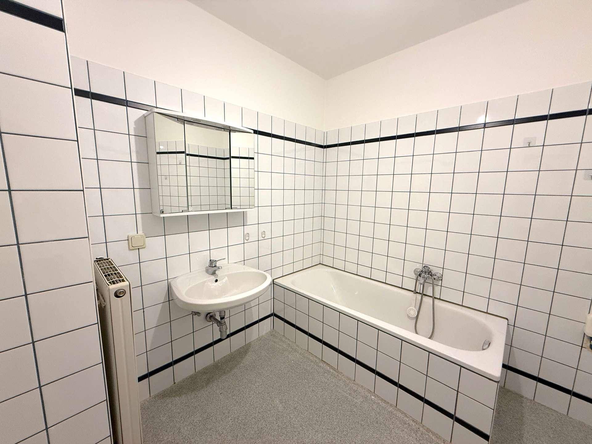 Badezimmer mit Badewanne, Waschbecken und weißen Fliesen mit schwarzem Dekorstreifen.
