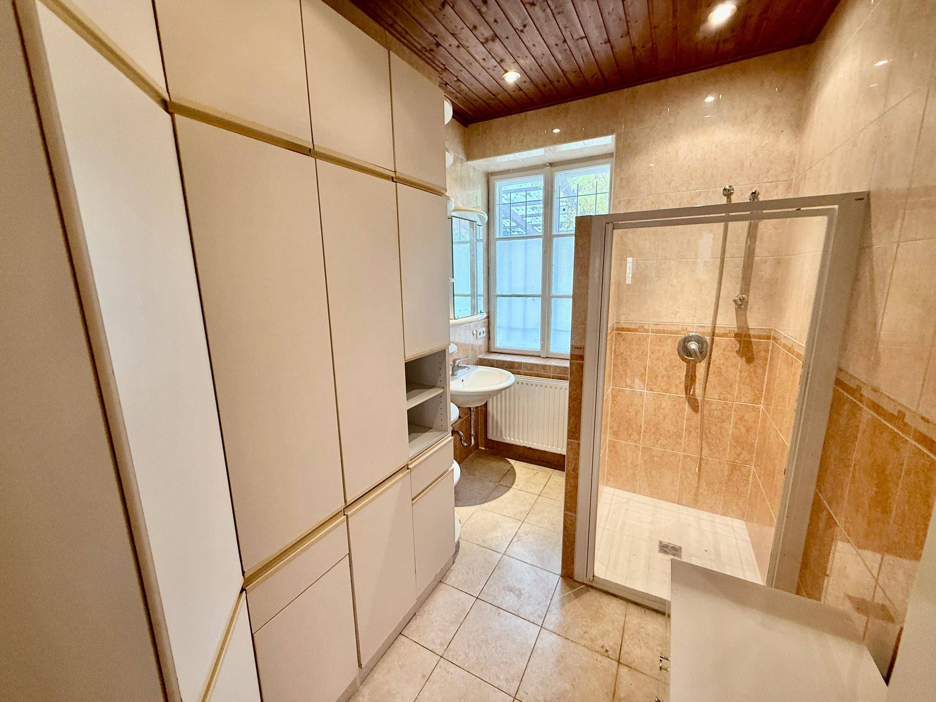 Badezimmer mit Duschkabine, Holzdecke und hellen Fliesen, inklusive Fenster für Tageslicht.