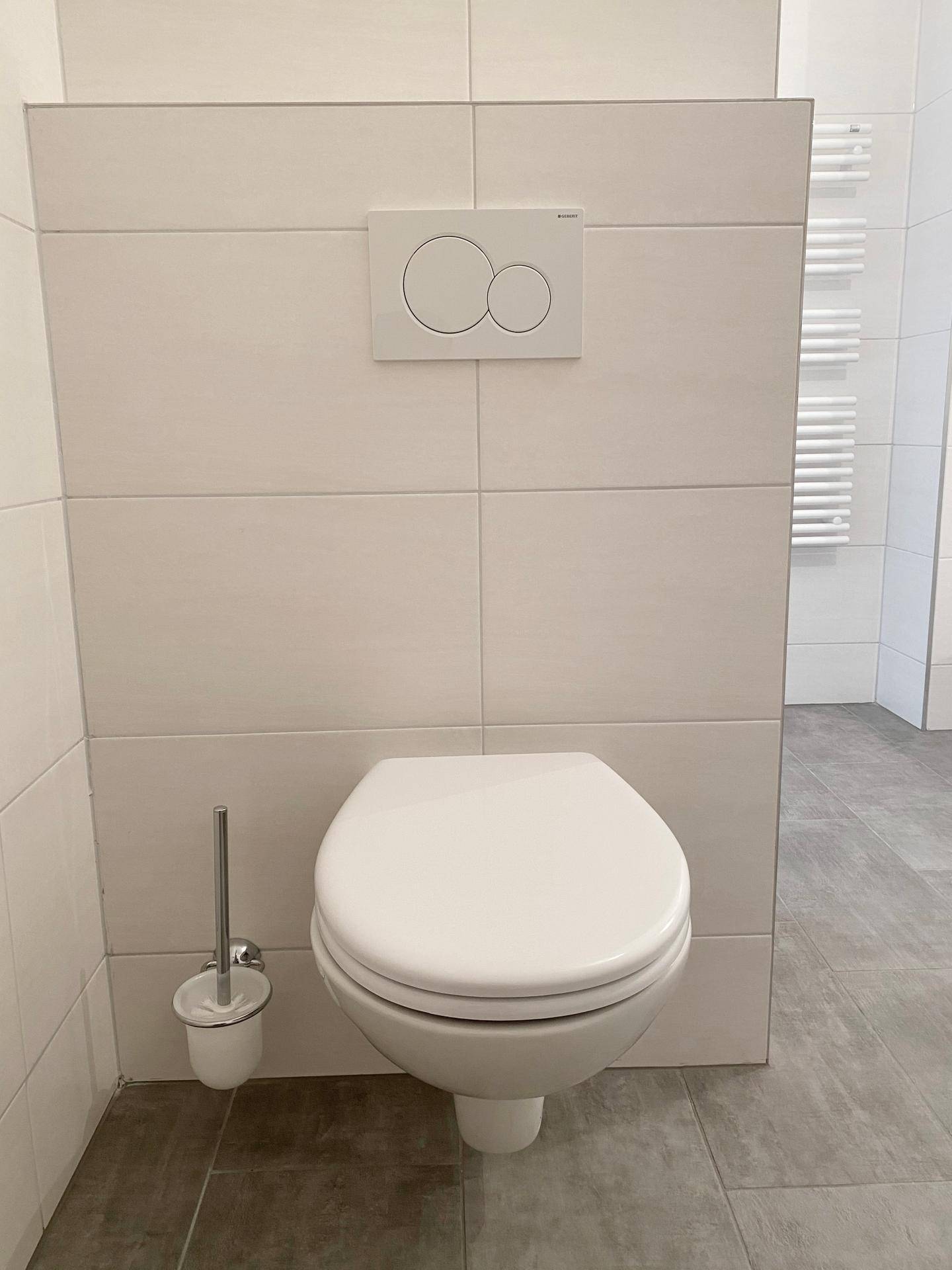 Modernes Wand-WC mit Spülkasten und weißem Bürstenhalter in einem gefliesten Raum.