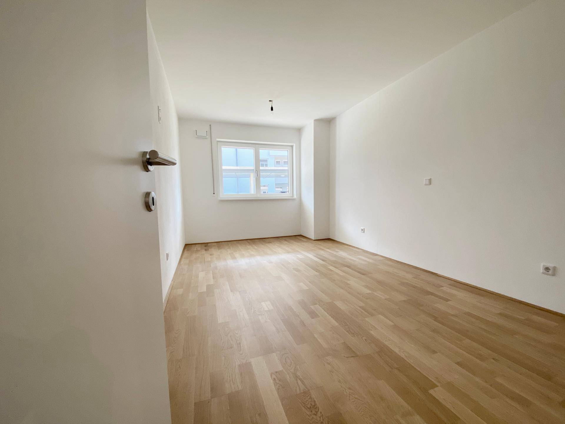 Helles, leeres Zimmer mit Holzfußboden und einem Fenster, bereit zur individuellen Gestaltung.