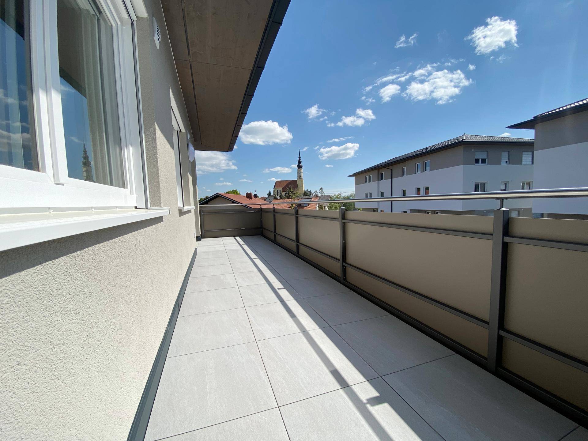 Langer, gefliester Balkon mit Metallgeländer und Blick auf die umliegende Bebauung und Kirche.