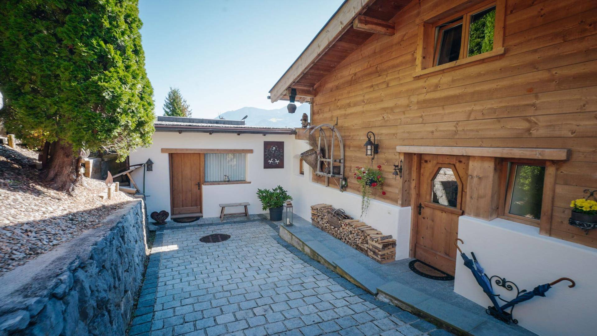 Gepflasterter Vorplatz mit Zugang zum Haus und Holzstapel an der Wand.