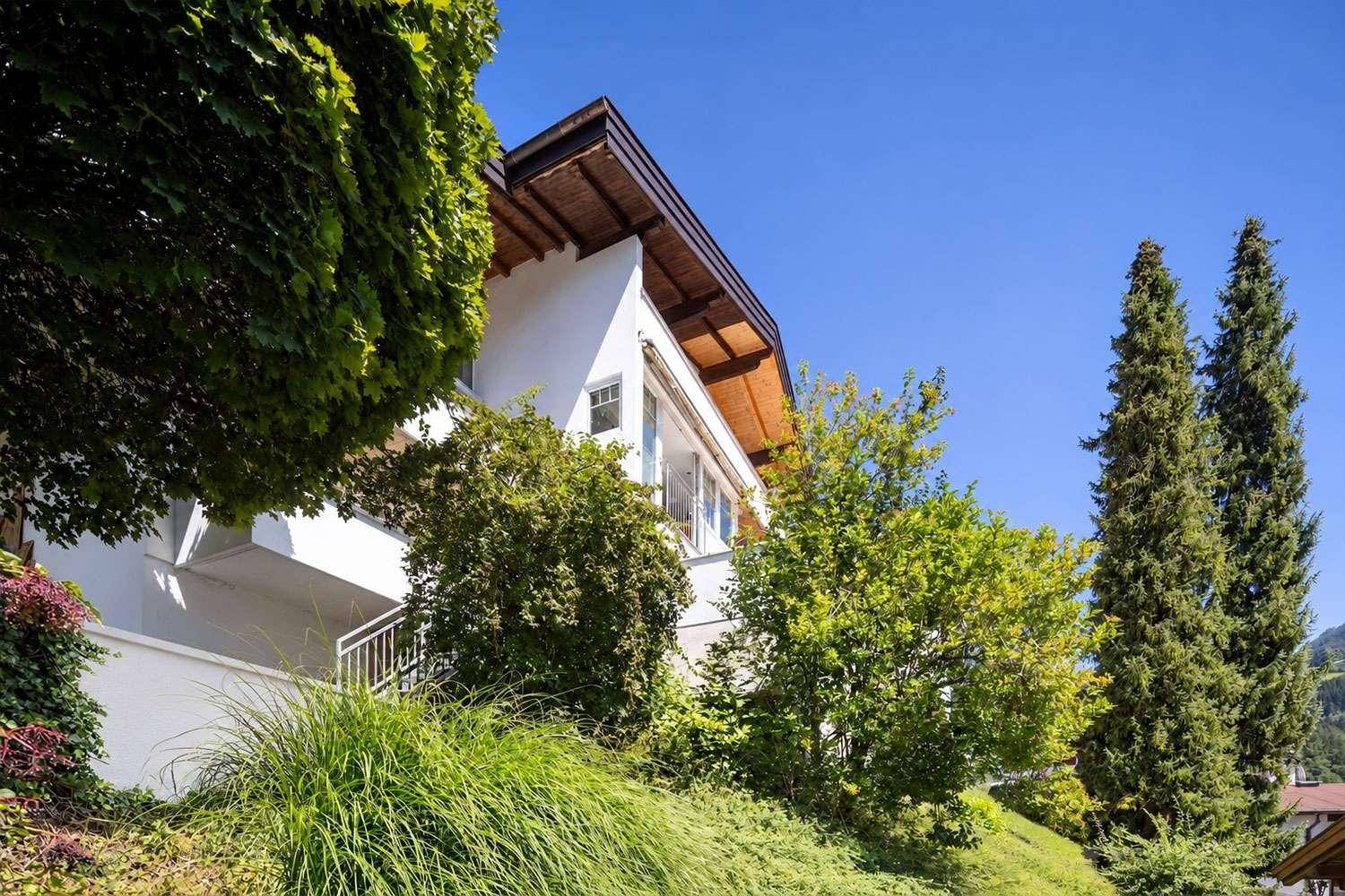 Seitenansicht des Hauses mit weißer Fassade, umgeben von dichtem Grün und hohen Bäumen.