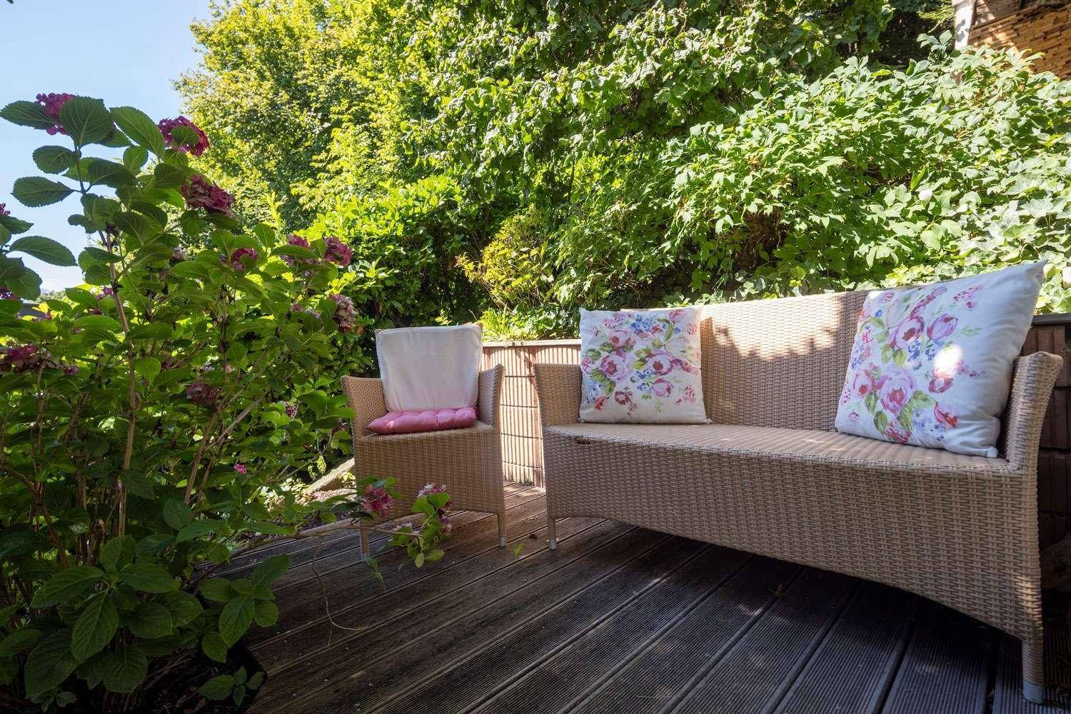 Gemütlicher Außenbereich auf einer Holzterrasse, umgeben von üppigem Grün und Blumen.