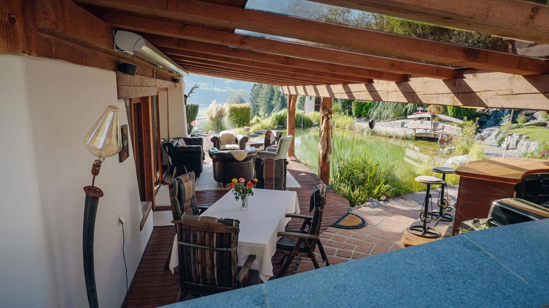 Überdachte Terrasse mit Esstisch, Lounge-Möbeln und Blick auf den Teich.