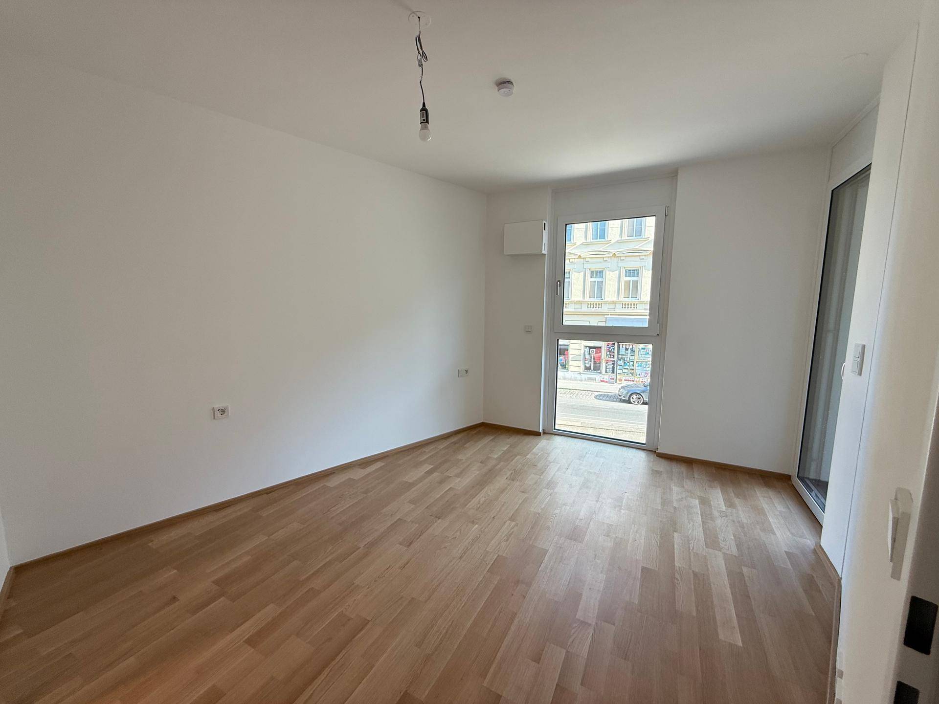 Leeres Zimmer mit Parkettboden und großem Fenster zur Straße.