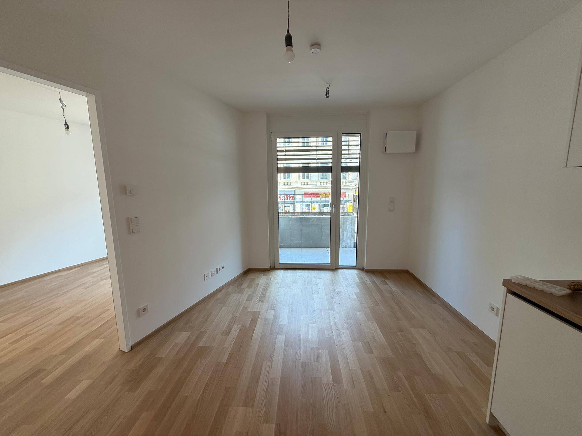 Helles, leeres Wohnzimmer mit Parkettboden und Zugang zu einem Balkon.