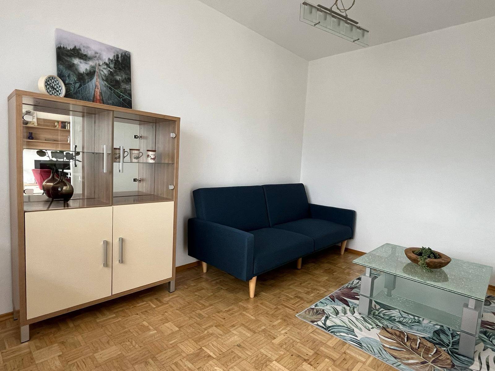 Gemütliches Wohnzimmer mit blauem Sofa, Glastisch und einem eleganten Vitrinenschrank.