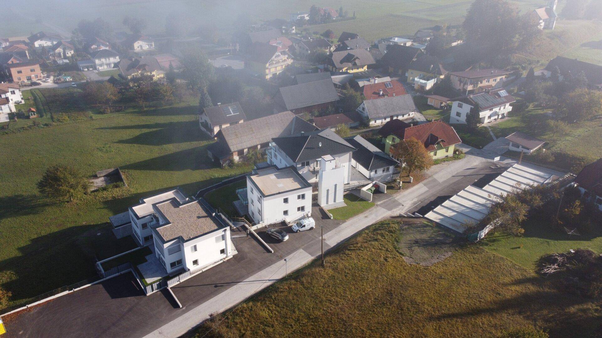 Luftaufnahme der Wohnanlage und des Dorfes, teilweise von Nebel bedeckt, in ländlicher Umgebung.