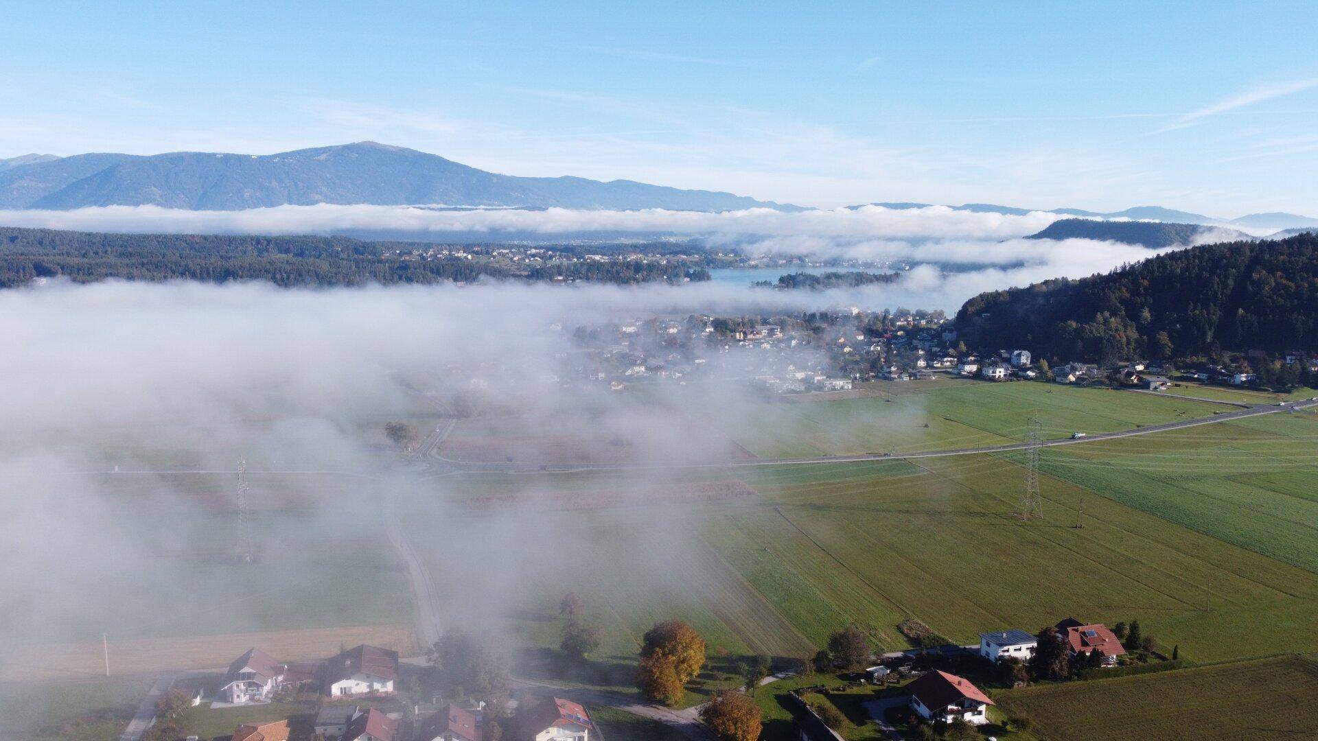 Weitläufige Luftaufnahme einer Stadt am See, umgeben von Nebel und Bergen.