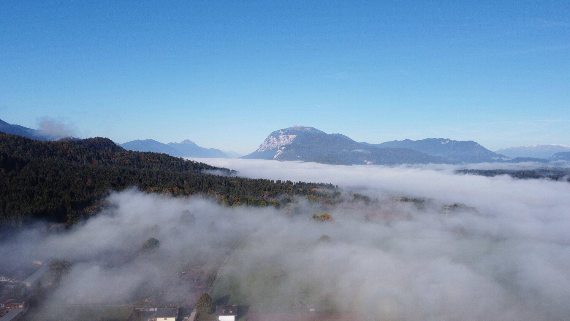 Panoramablick aus der Luft über eine nebelverhangene Landschaft mit majestätischen Bergen im Hintergrund.