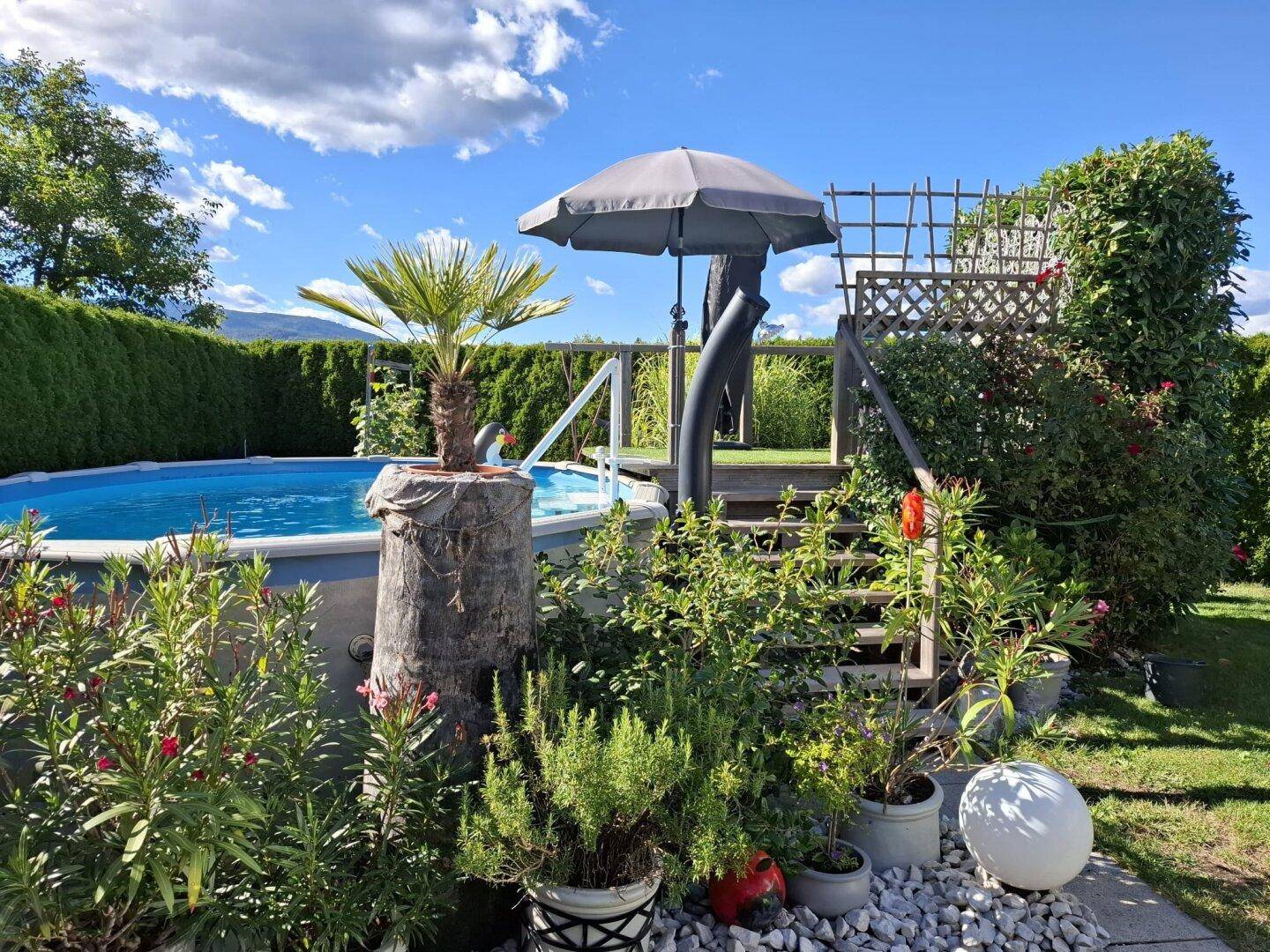 Gartenbereich mit Aufstellpool, Sonnenschirm und üppiger Bepflanzung unter strahlend blauem Himmel.