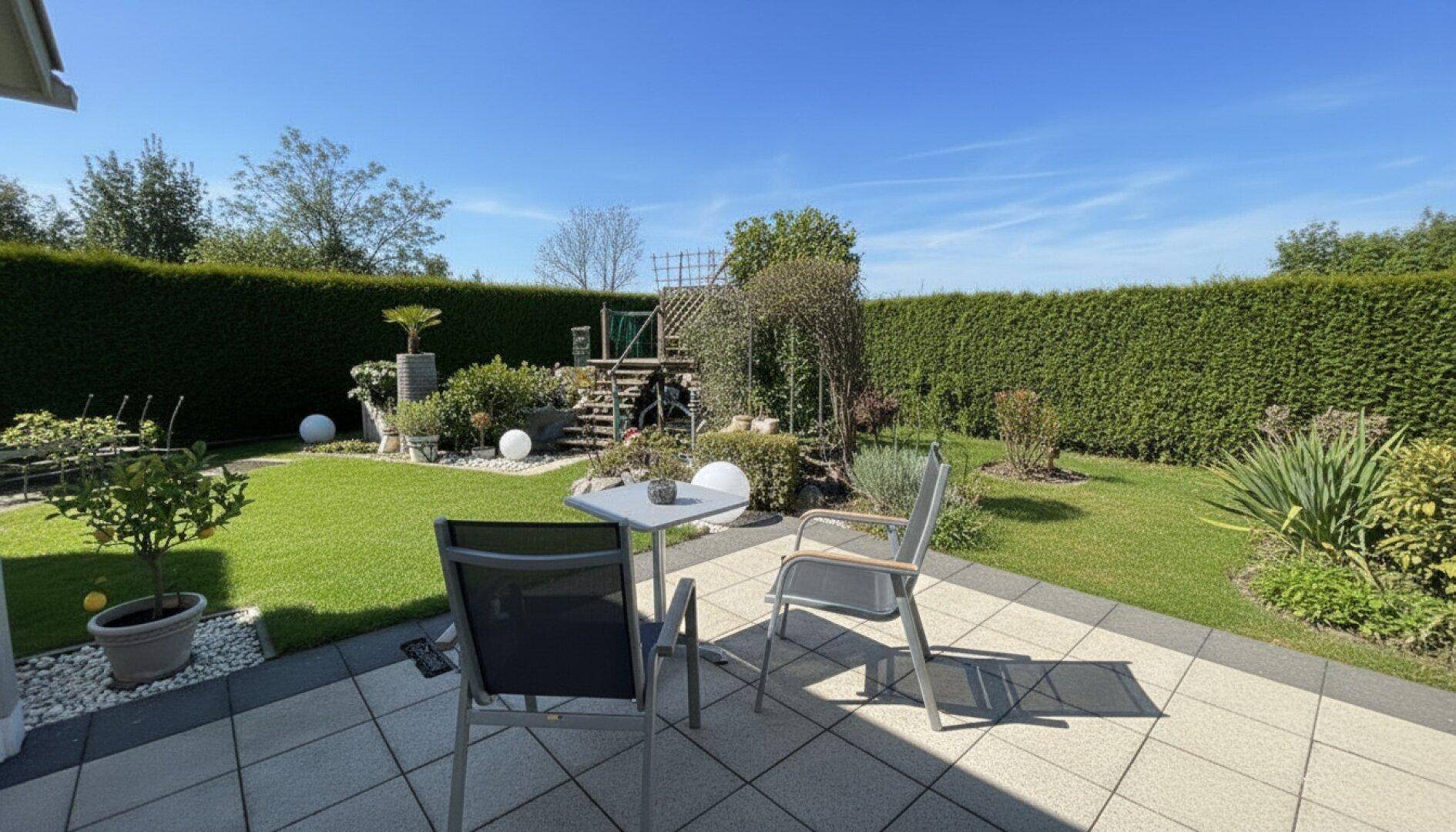Geräumige Terrasse mit Gartenmöbeln und gepflegtem Rasen, umgeben von einer hohen Hecke.