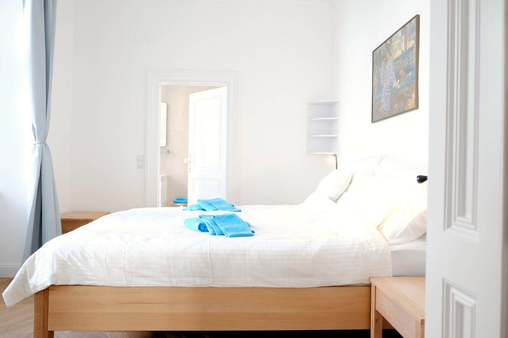 Helles Schlafzimmer mit Doppelbett, Nachttischen und Zugang zu einem weiteren Raum.