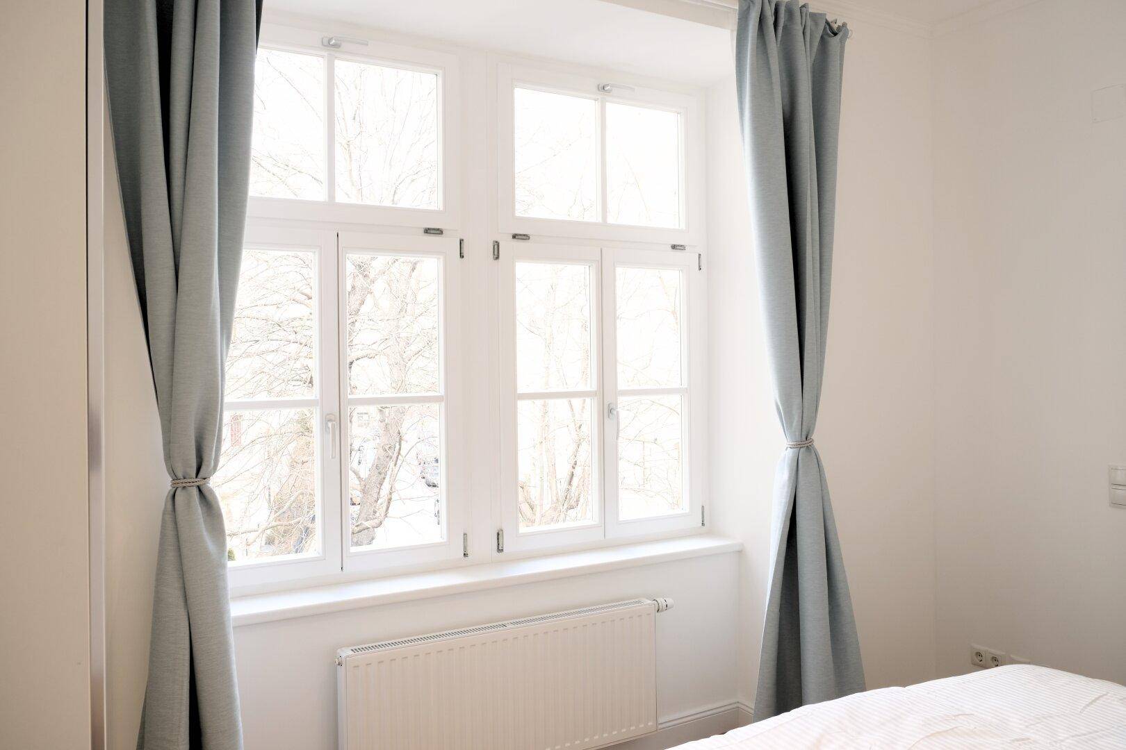 Großes Fenster im Schlafzimmer mit hellblauen Vorhängen und Heizkörper darunter.