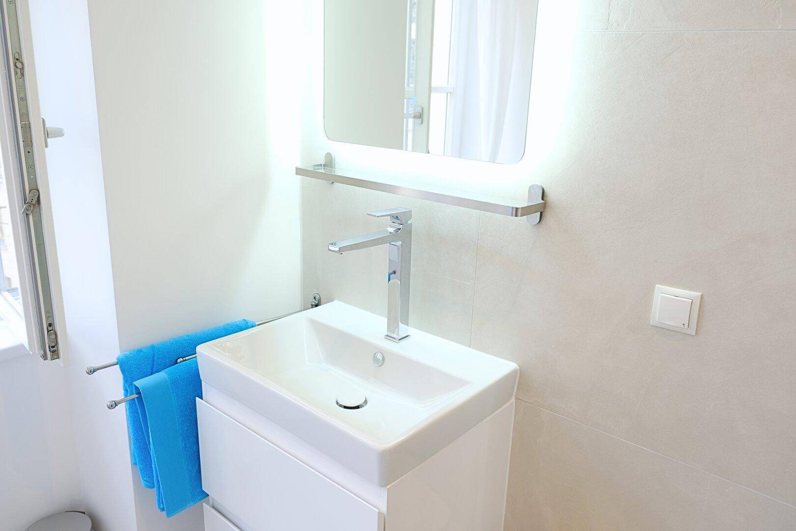 Modernes Badezimmer mit Einzelwaschbecken, beleuchtetem Spiegel und hellen Fliesen.