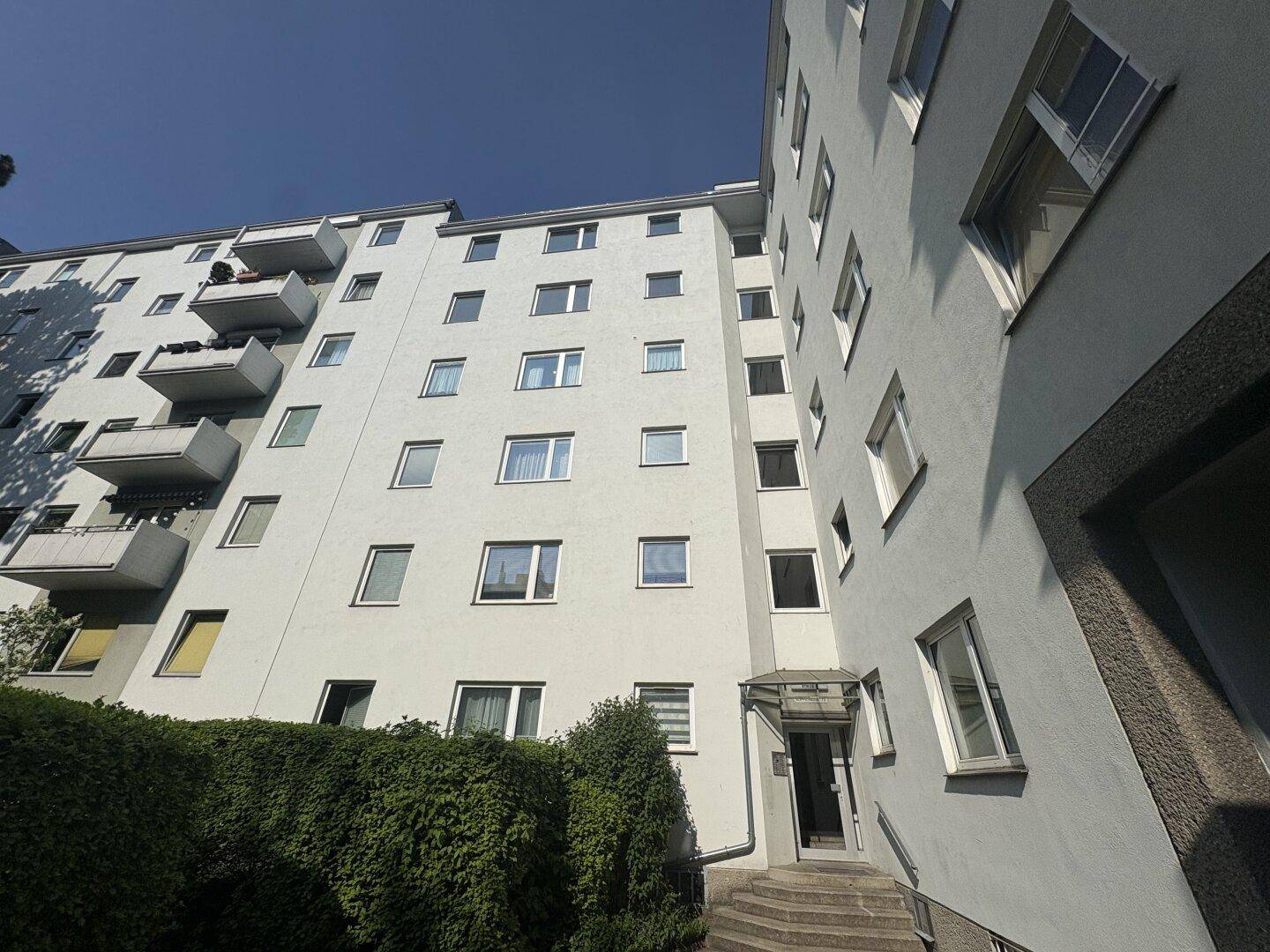 Fassade eines Mehrfamilienhauses mit Balkonen und vielen Fenstern unter blauem Himmel, solide Bauweise.