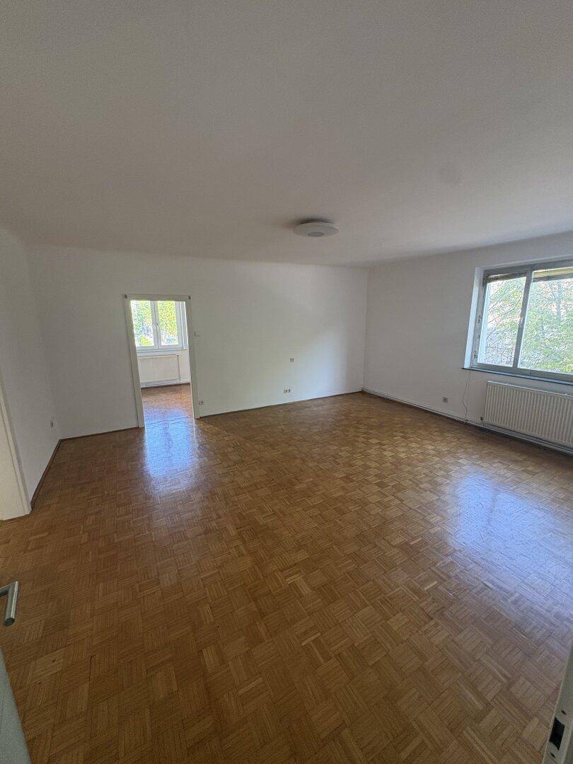 Geräumiges Wohnzimmer mit Parkettboden und Blick ins Grüne durch das Fenster, bereit zur individuellen Gestaltung.
