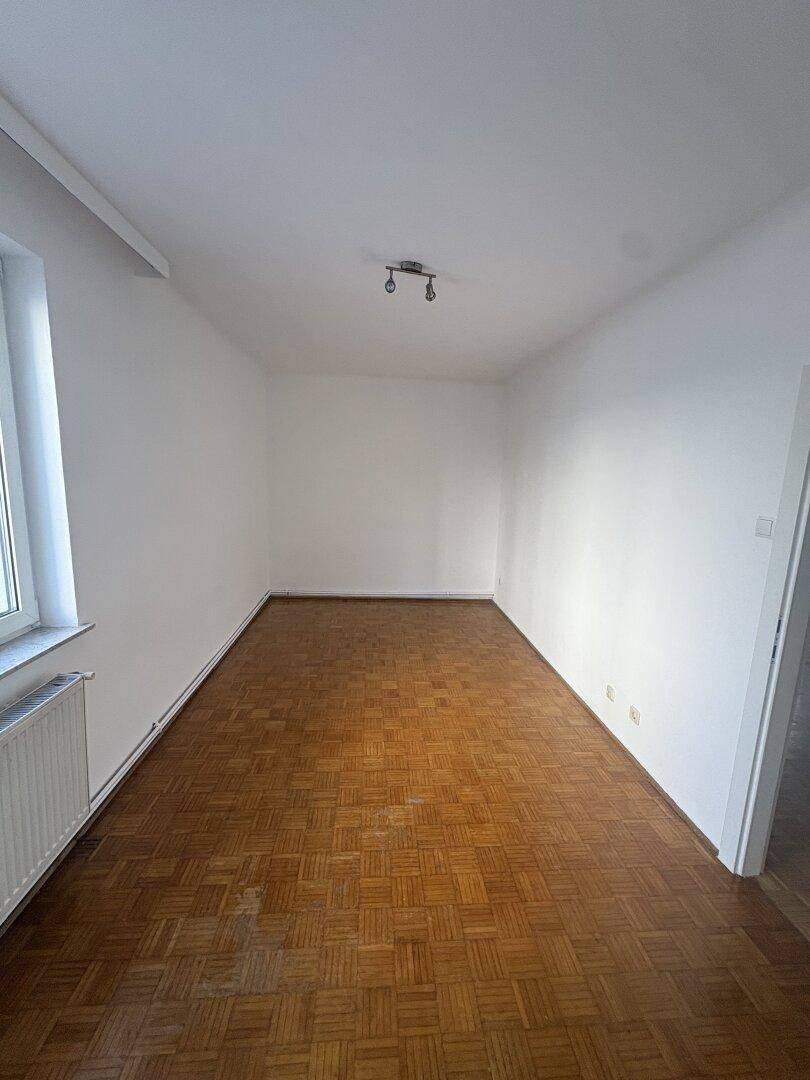 Leeres Zimmer mit Parkettboden, weißen Wänden und einem Fenster für Tageslicht, vielseitig nutzbar.